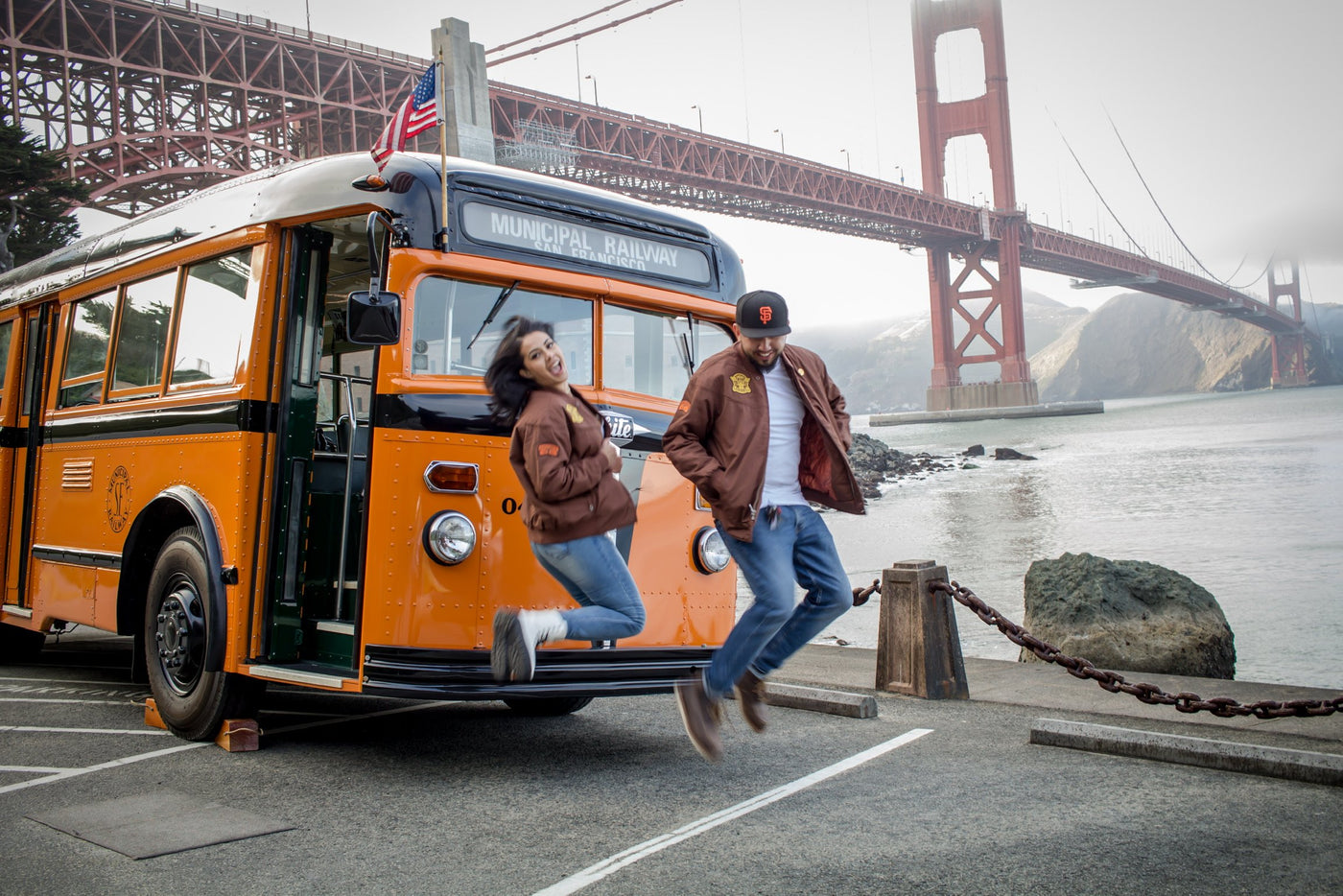 Couple jumping in front of San Francisco Golden Gate Bridge and orange bus in brown Jeremy Fish collaboration brown Derby Jackets