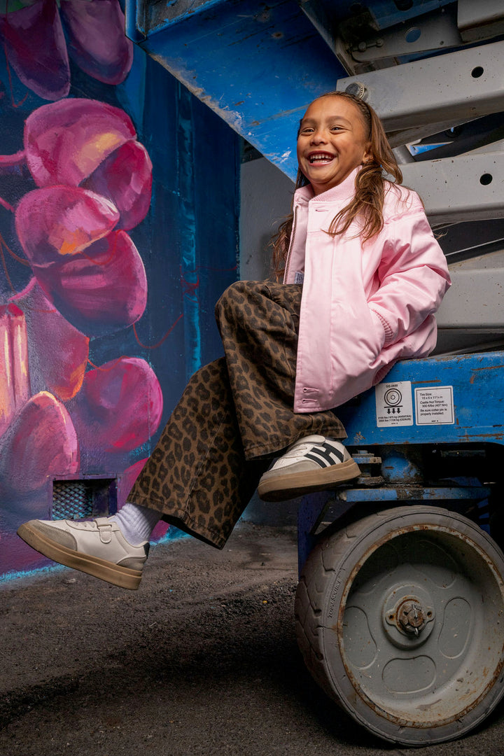 A young girl with a smile on her face sits on the blue wheel of a construction vehicle, wearing a light pink puffer jacket, leopard-print pants, and white sneakers. To her left is a colorful mural painted on the side of the vehicle.