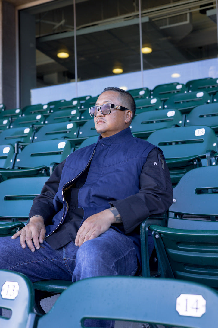 A man wearing a navy blue vest and dark shirt sits in green stadium seating, looking off-camera. The background shows rows of green stadium seats and a modern building interior.
