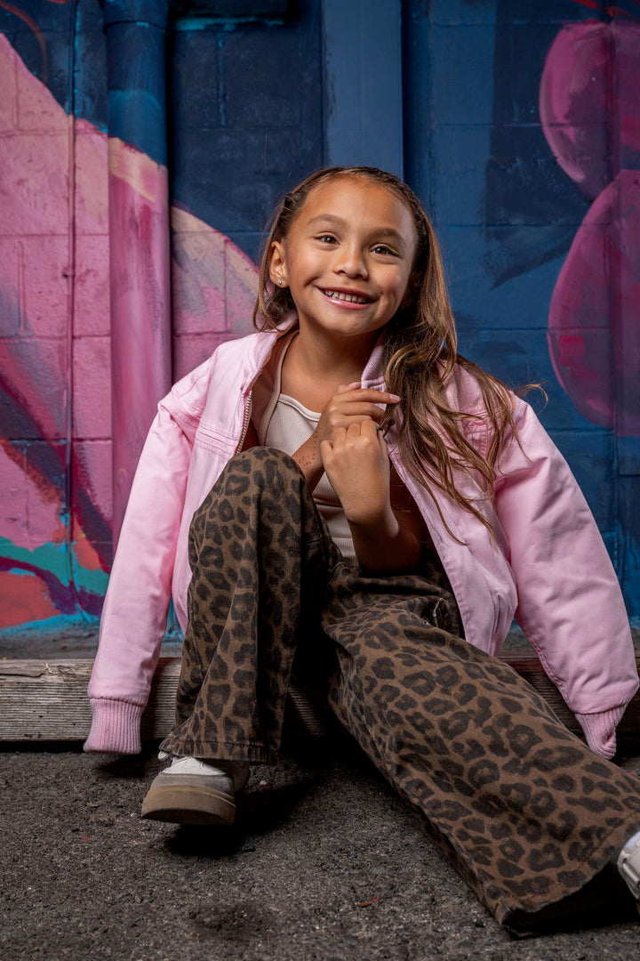 A young girl with long brown hair sits on a dark asphalt ground in front of a colorful graffiti wall. She is wearing a light pink bomber jacket over a white t-shirt and brown leopard-print wide-leg pants.
