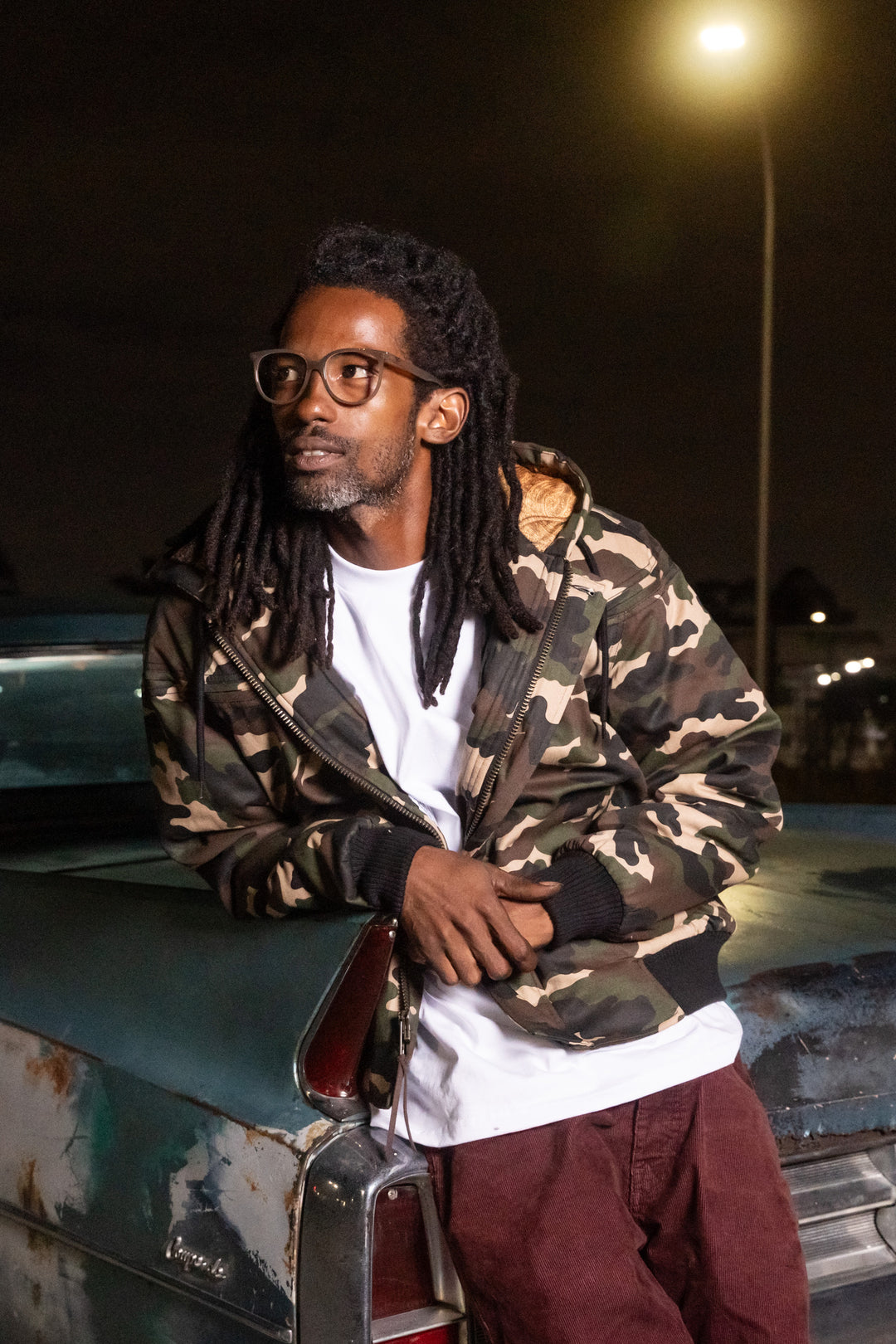 Man with dreadlocks wearing a camouflage jacket and glasses, sitting on a vehicle at night.