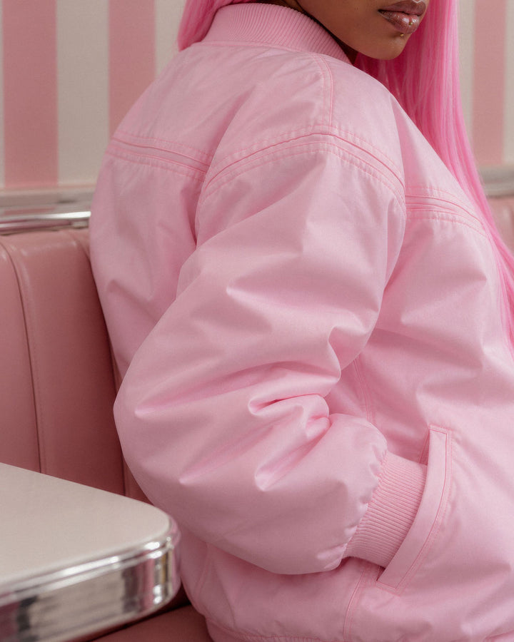 A close-up of a person wearing a light pink satin bomber jacket, seated in a retro-style pink booth with pink striped walls.