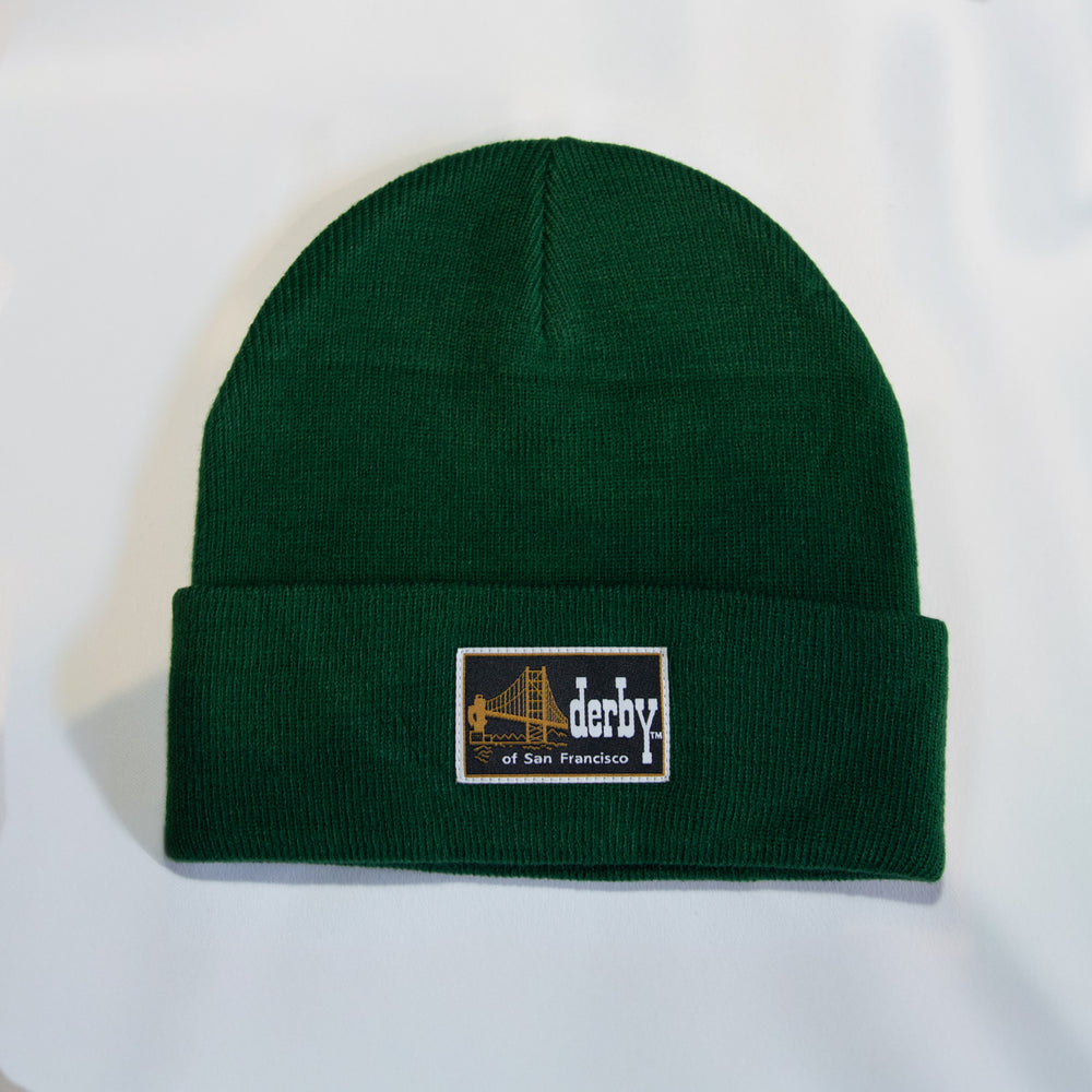 A dark green ribbed knit beanie hat with a folded cuff, featuring a rectangular embroidered patch on the front that reads 'derby of San Francisco' with a bridge graphic.