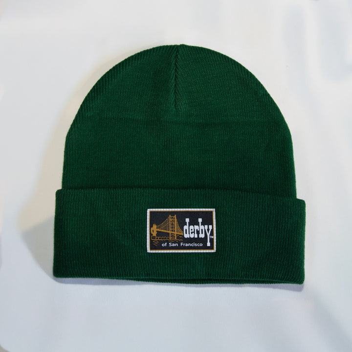 A dark green ribbed knit beanie hat with a folded cuff, featuring a rectangular embroidered patch on the front that reads &