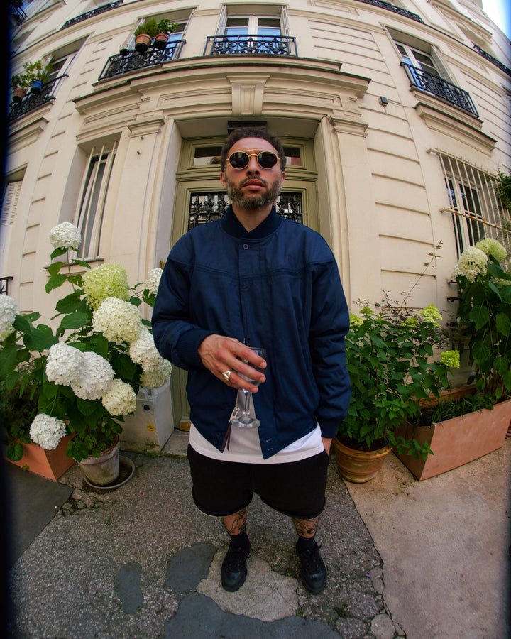 Man in sunglasses and a blue jacket standing in front of a building with flowers and plants.