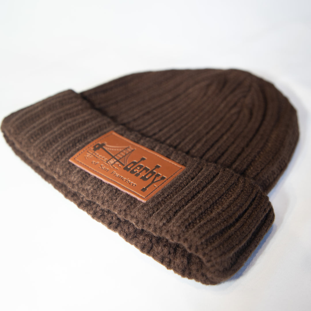 A dark brown ribbed knit beanie hat featuring a brown leather patch on the front cuff with the brand name &