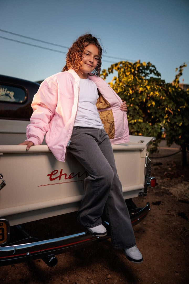 A young woman with curly brown hair leans against the tailgate of a vintage cream pickup truck outdoors. She is wearing a light pink bomber jacket over a white t-shirt, gray wide-leg jeans, and white sneakers.