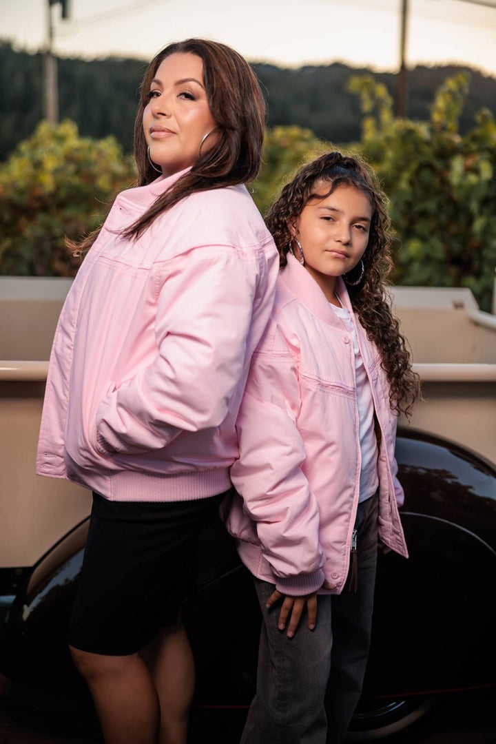 A woman and a young girl standing back-to-back in matching light pink bomber jackets, photographed outdoors with a car and greenery in the background.