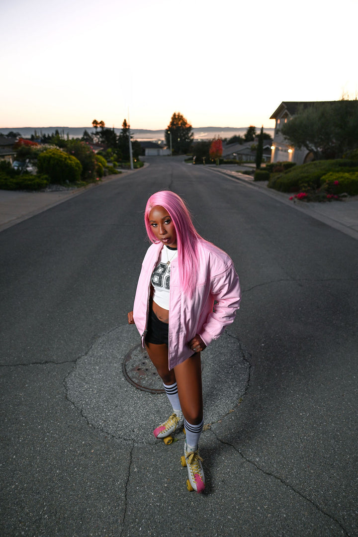 A person standing on a suburban street at sunset wearing a light pink oversized jacket, a white cropped graphic T-shirt, black shorts, white knee-high socks, and pink-and-yellow sneakers.