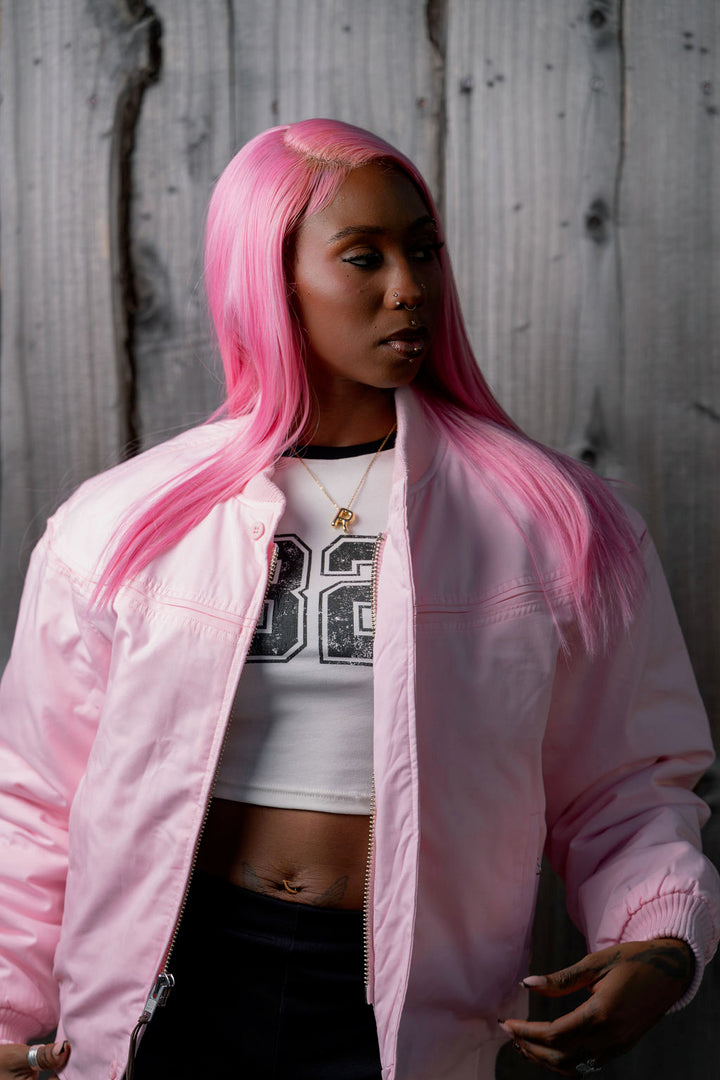 A model with vibrant pink hair wearing a light pink bomber jacket over a white graphic crop top, standing against a rustic gray wooden wall.