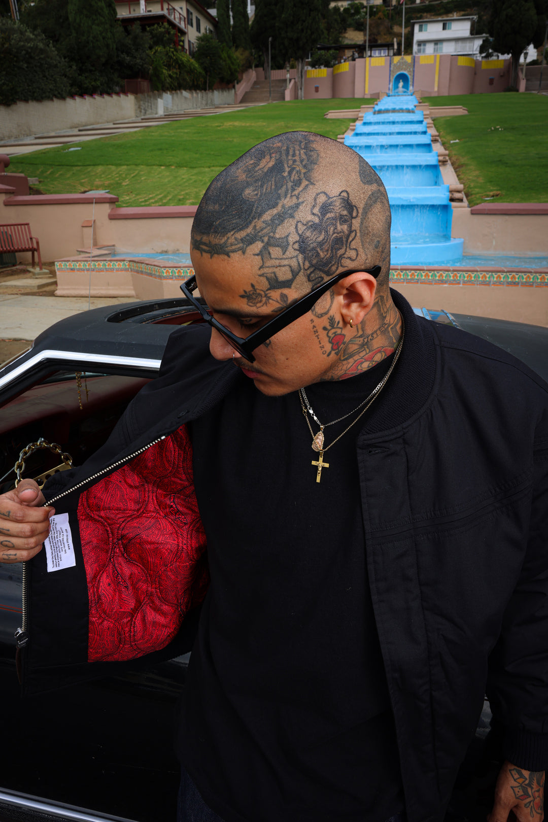 A tattooed man wearing black sunglasses, a black shirt, and a black jacket stands by an open car door, holding up a red and black patterned fabric item. A blue water feature and green lawn are visible in the background.