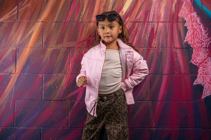 A young girl stands in front of a colorful painted brick wall wearing a light pink zip-up jacket over a cream t-shirt, leopard-print pants, and black sunglasses on her head.