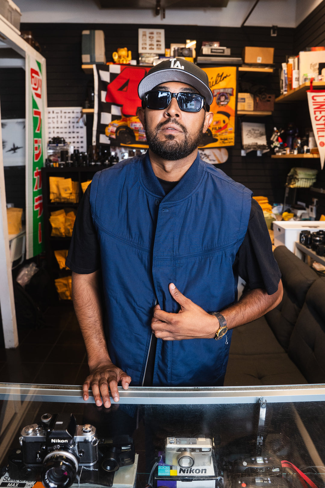 A man wearing a navy blue zip-up vest over a black t-shirt, a gray baseball cap, and sunglasses, standing in a cluttered vintage camera shop.