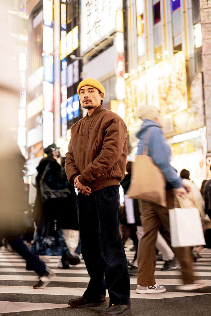 Man in a brown sweater and yellow beanie standing on a busy street with neon lights in the background