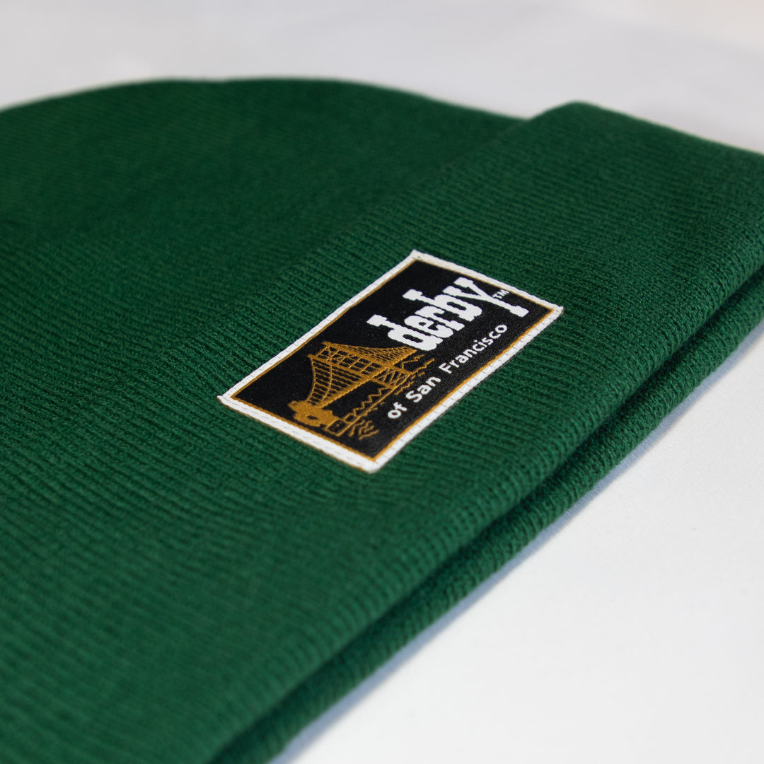 A close-up of a dark green ribbed knit beanie with a rectangular embroidered patch on the cuff. The patch is black with a gold outline of the Golden Gate Bridge and white text reading 'denby of San Francisco'.