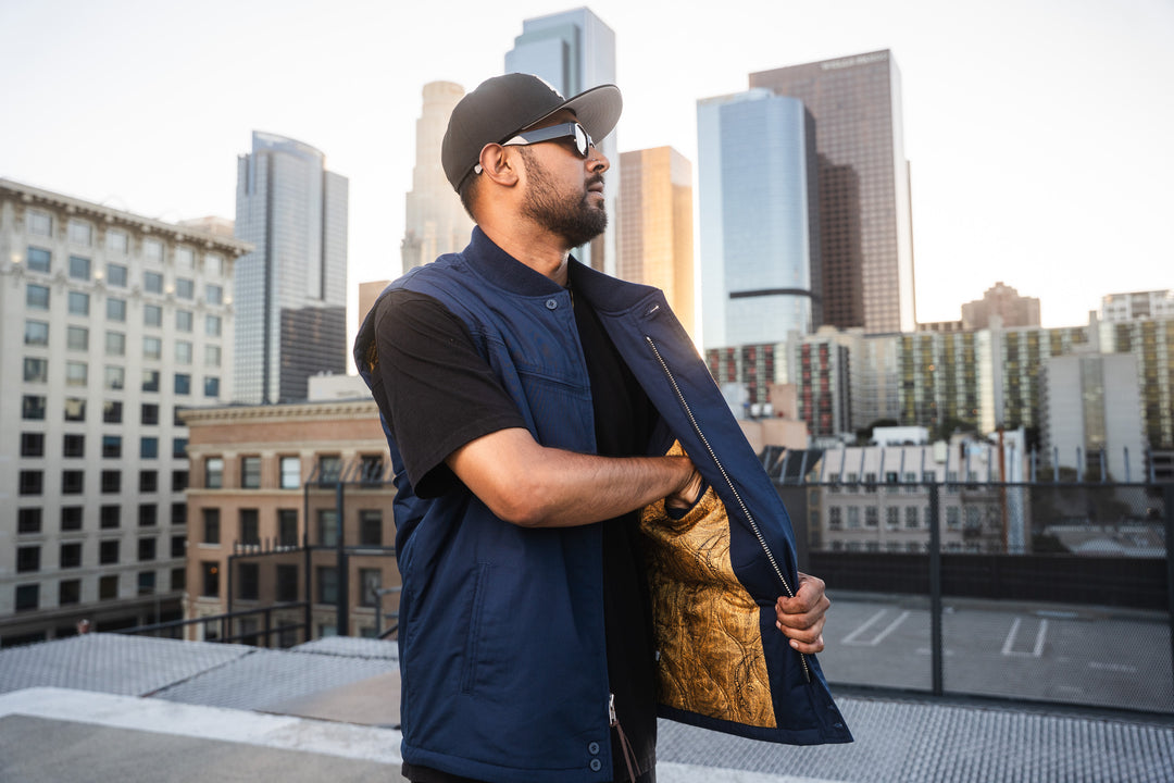 A man wearing a navy blue zippered vest over a black t-shirt stands on a rooftop with a city skyline in the background, holding open the vest to show a gold patterned lining.