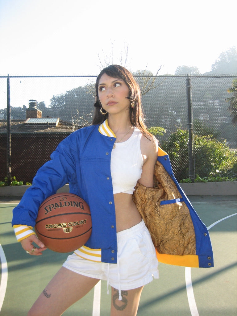 Person holding a basketball on a court wearing a blue and gold jacket.