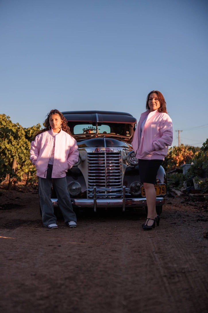 Two women standing on a dirt road in front of a vintage black car, wearing matching light pink jackets. The scene is set outdoors under a clear blue sky.