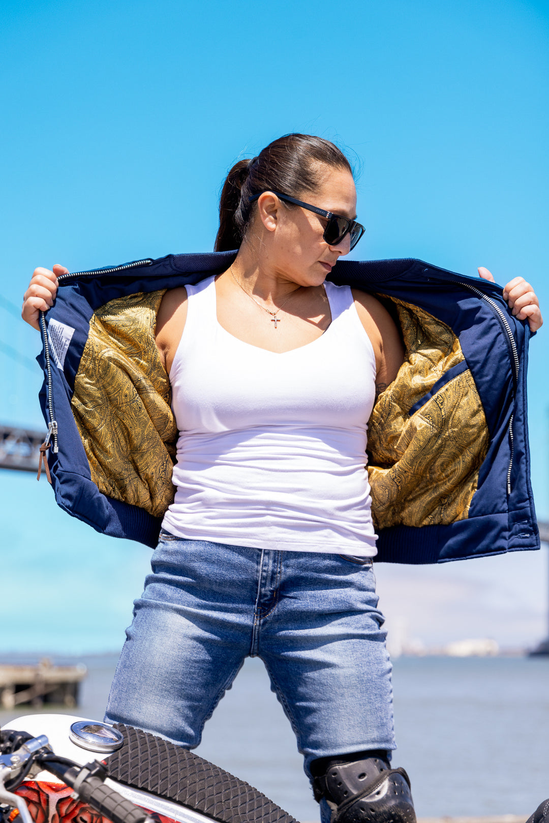 Woman wearing a gold and navy jacket with a white tank top and blue jeans, standing on a beach.