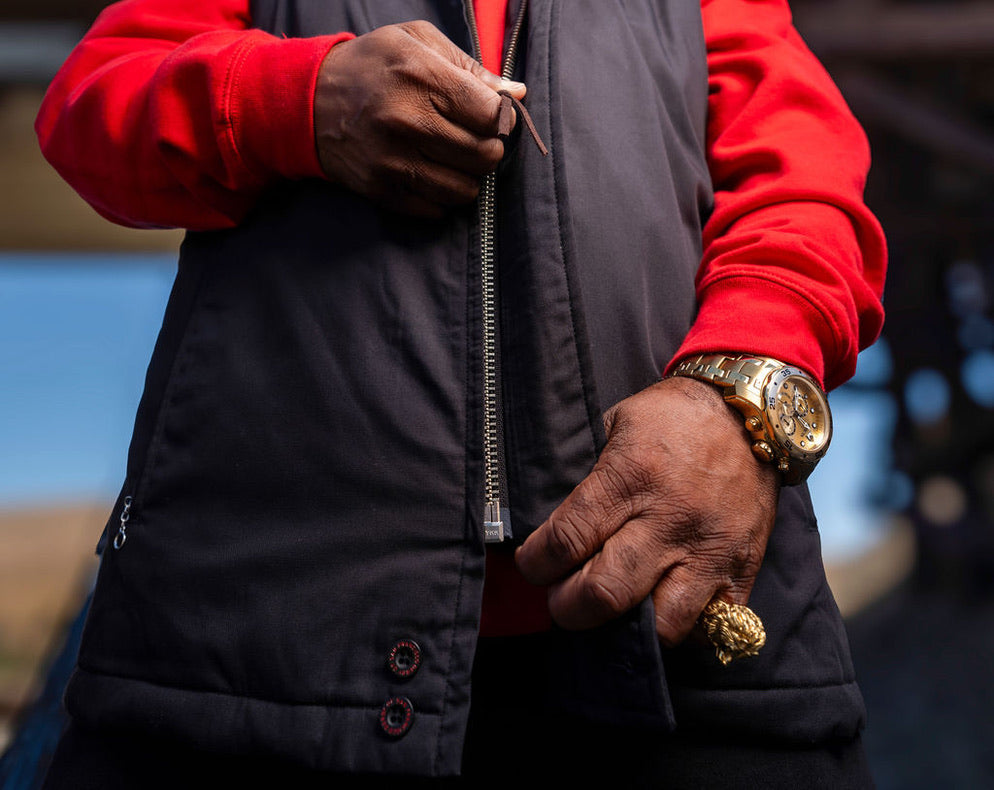 Person wearing a red jacket and black vest with gold jewelry, blurred background
