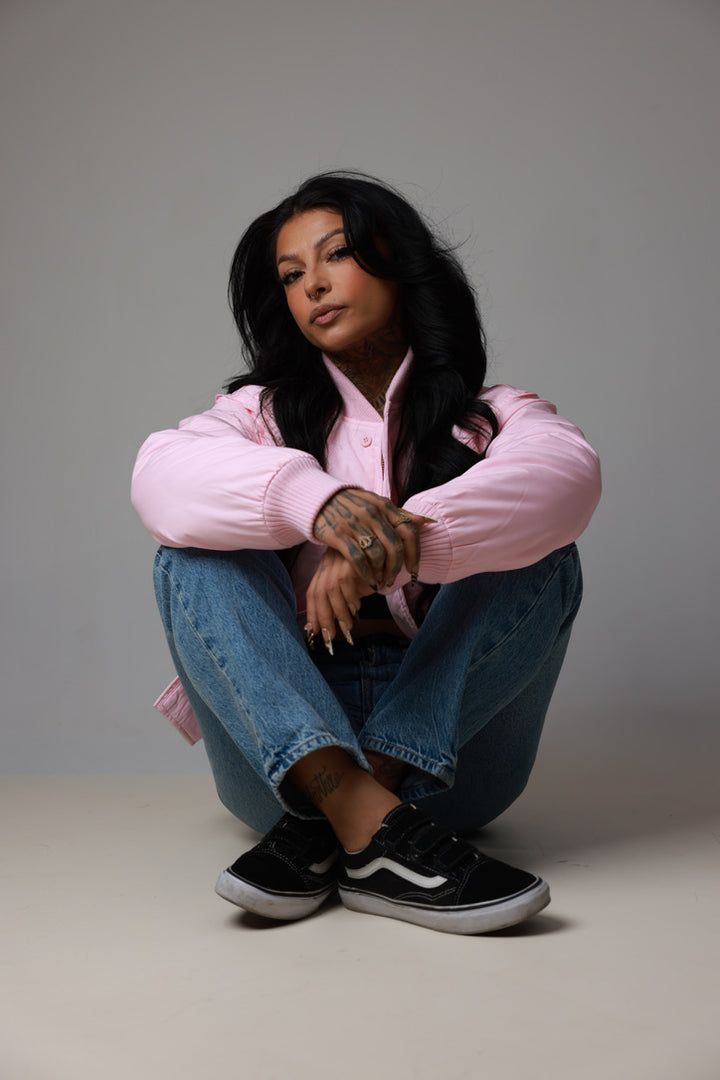 A woman sits cross-legged on a light gray studio floor wearing a light pink bomber jacket, blue jeans, and black Vans sneakers with white side stripes.