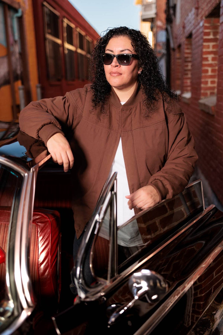 Person wearing sunglasses and a brown jacket standing next to a vintage car.
