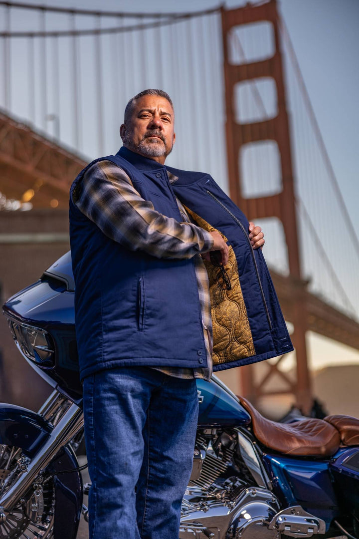 A man wearing a navy blue quilted motorcycle vest over a plaid shirt and jeans stands next to a blue motorcycle with the Golden Gate Bridge in the background.