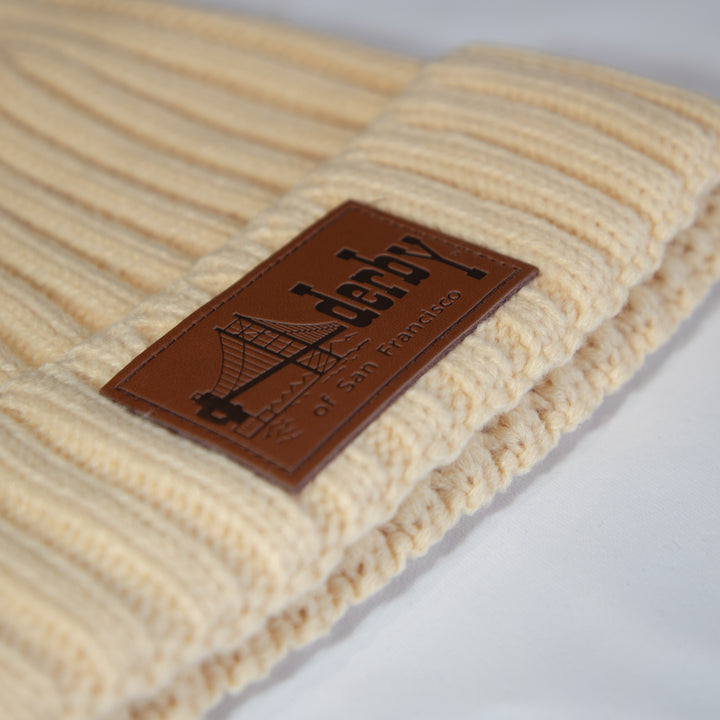 Close-up of a cream ribbed knit beanie with a brown rectangular leather patch reading Derby of San Francisco, featuring a bridge graphic.