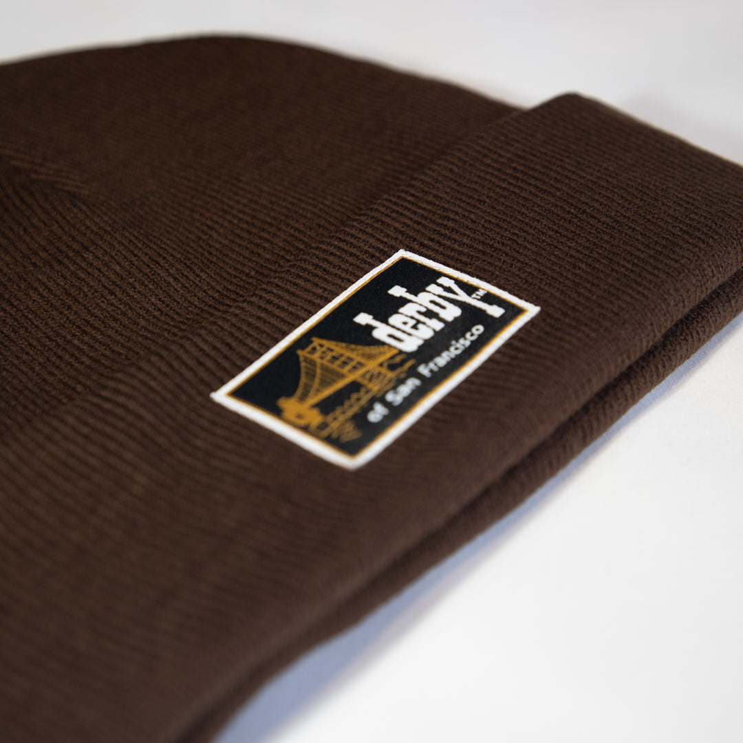 A close-up of a dark brown ribbed knit beanie with a rectangular embroidered patch on the cuff. The patch is black with a gold bridge graphic and white text reading &
