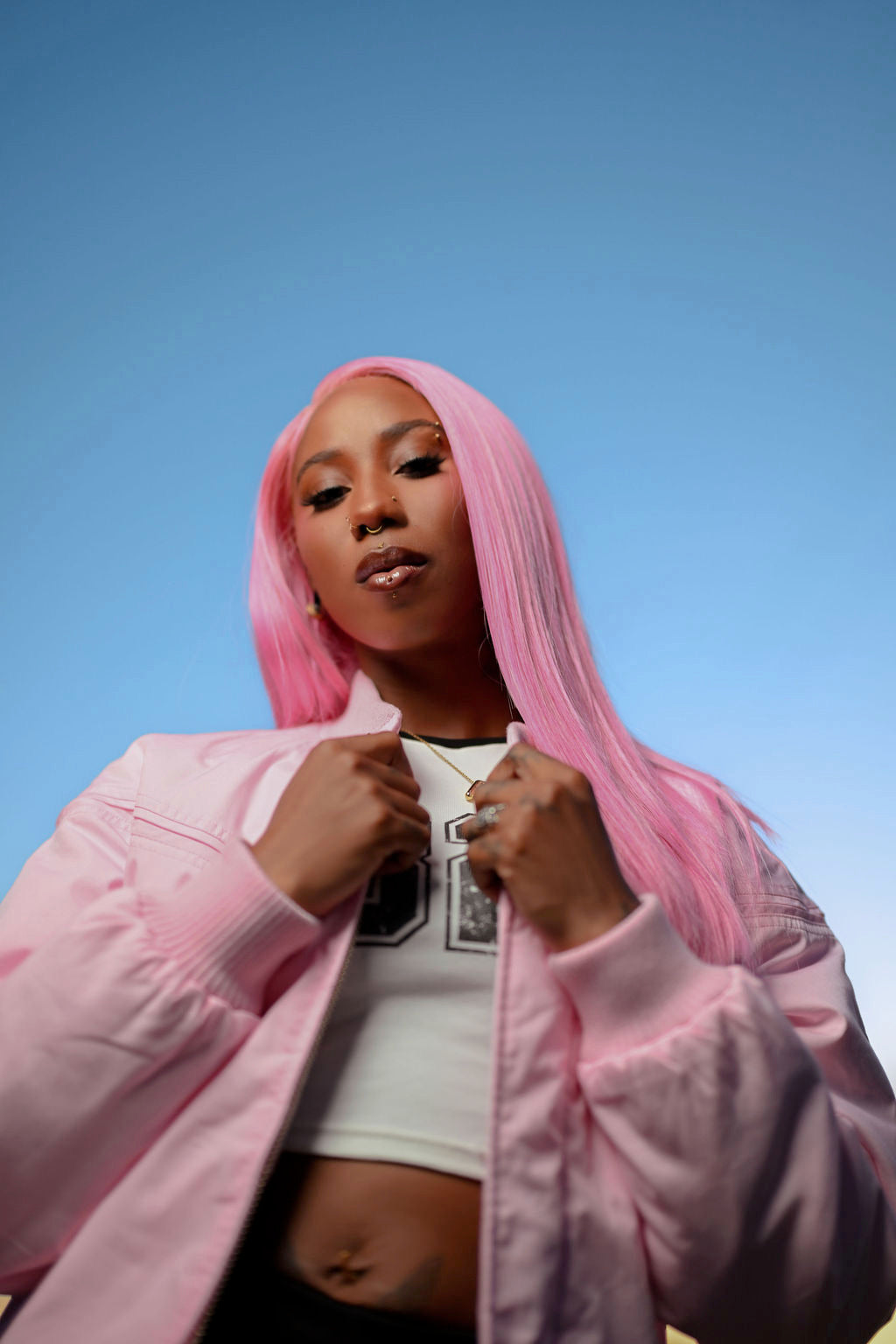 A woman with long pink hair wearing a light pink bomber jacket over a white crop top, photographed against a clear blue sky.