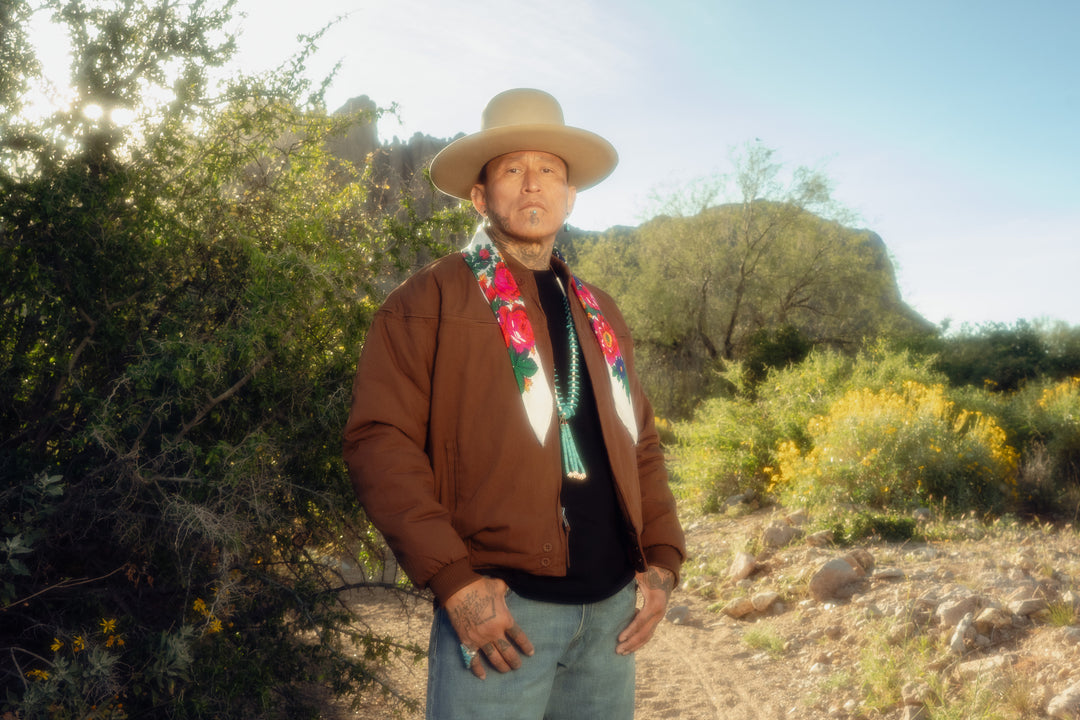 Man in a brown jacket and hat standing in a desert landscape with greenery and clear sky.