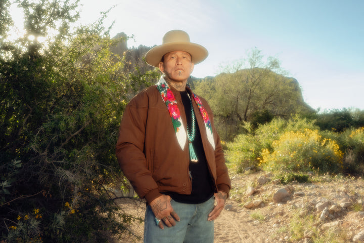 Man in a brown jacket and hat standing in a desert landscape with greenery and clear sky.