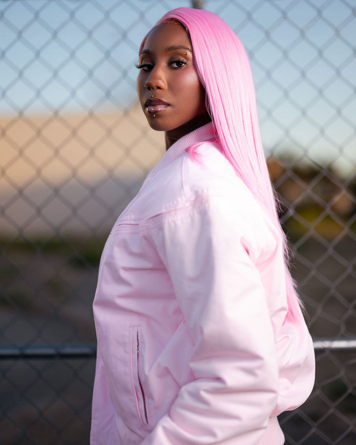A woman with long, straight bright pink hair is shown in profile wearing a light pink jacket, standing in front of a chain-link fence.