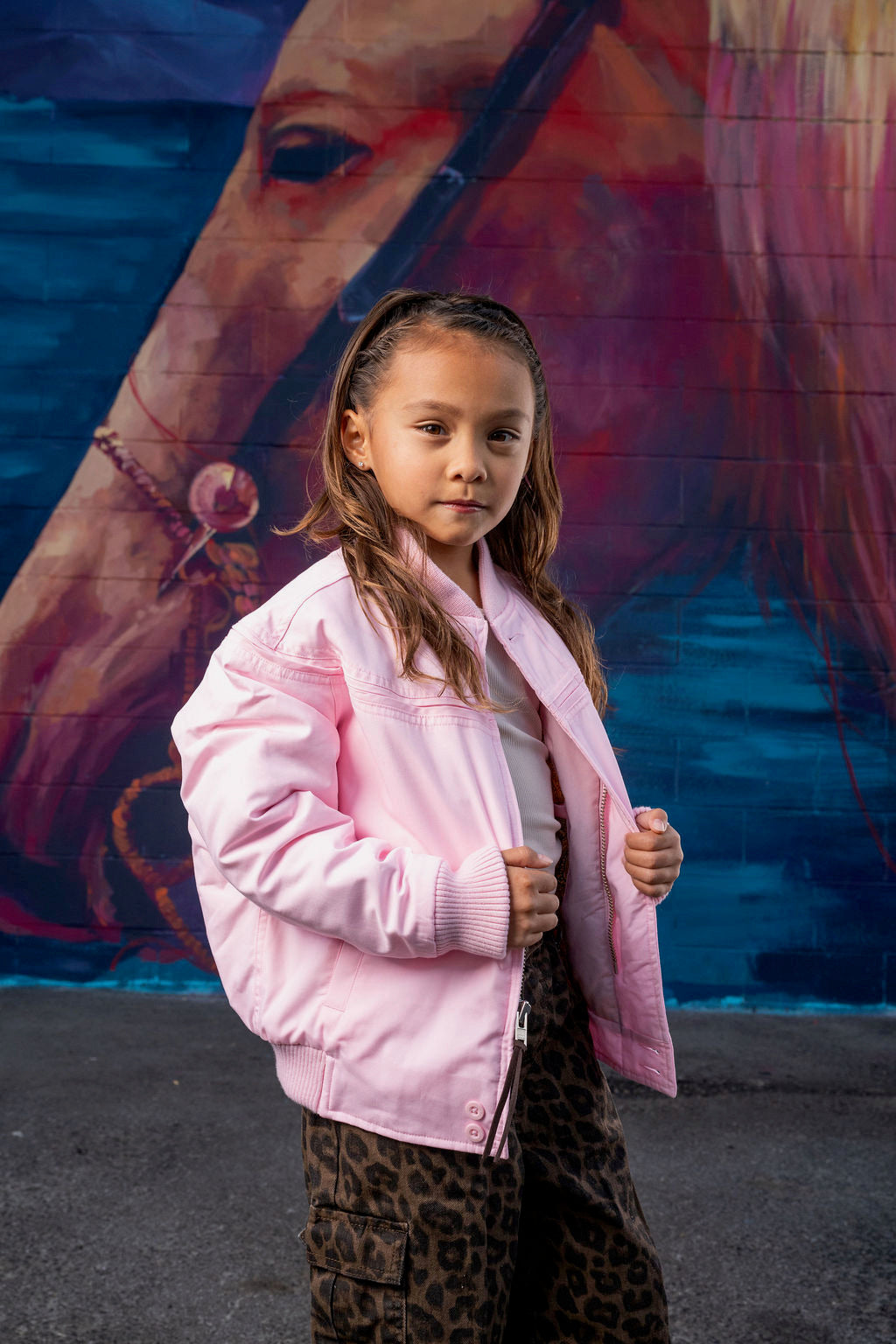 A young girl stands in front of a colorful mural wearing a light pink bomber jacket, a white top, and leopard-print cargo pants.