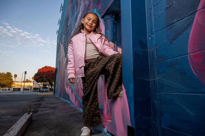 A young girl wearing a light pink puffer jacket, beige top, and leopard-print pants leans against a blue and pink graffiti wall outdoors.