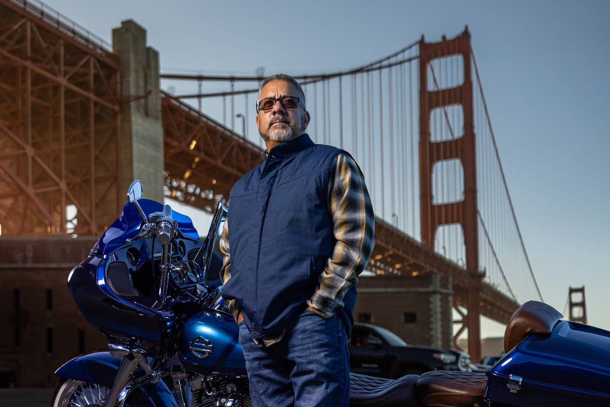 A man wearing sunglasses, a navy vest over a plaid shirt, and jeans stands next to a blue motorcycle in front of the Golden Gate Bridge.