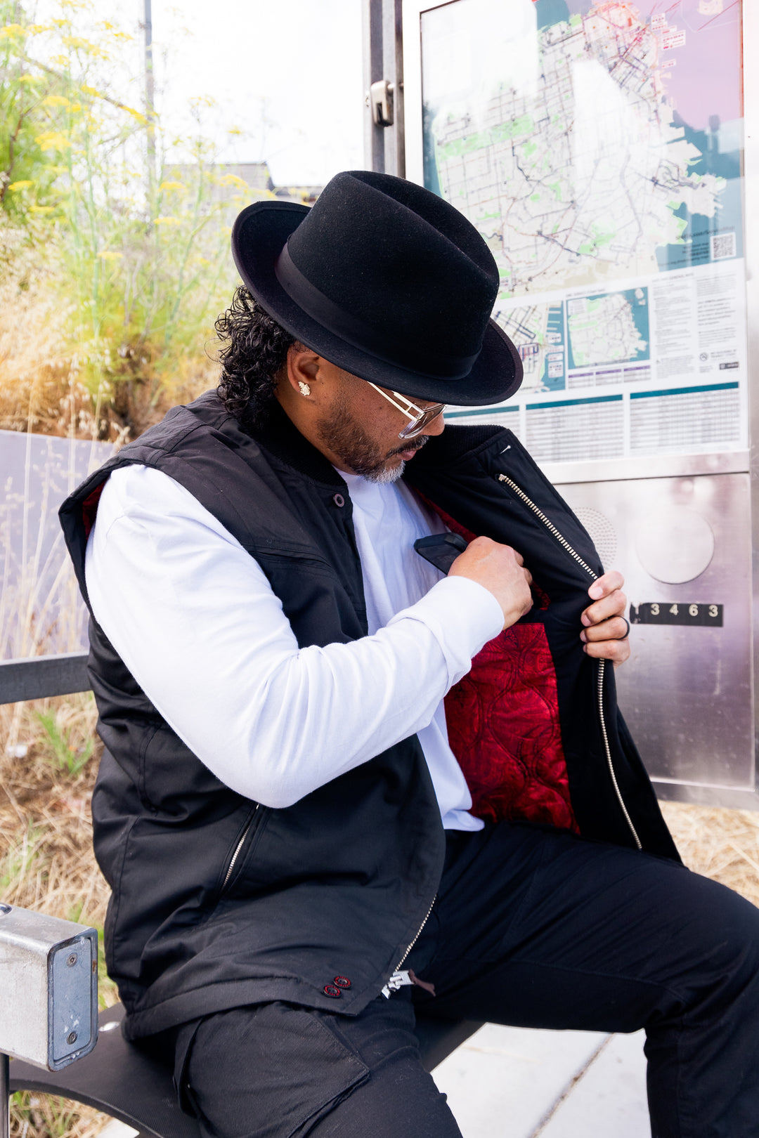 Man sitting on a bench wearing a black hat, white shirt, and black vest with a red interior.