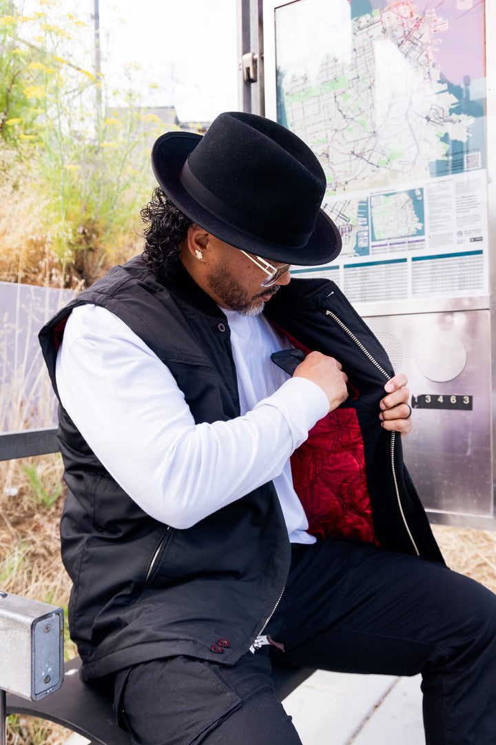 Man sitting on a bench wearing a black hat, white shirt, and black vest with a red interior.