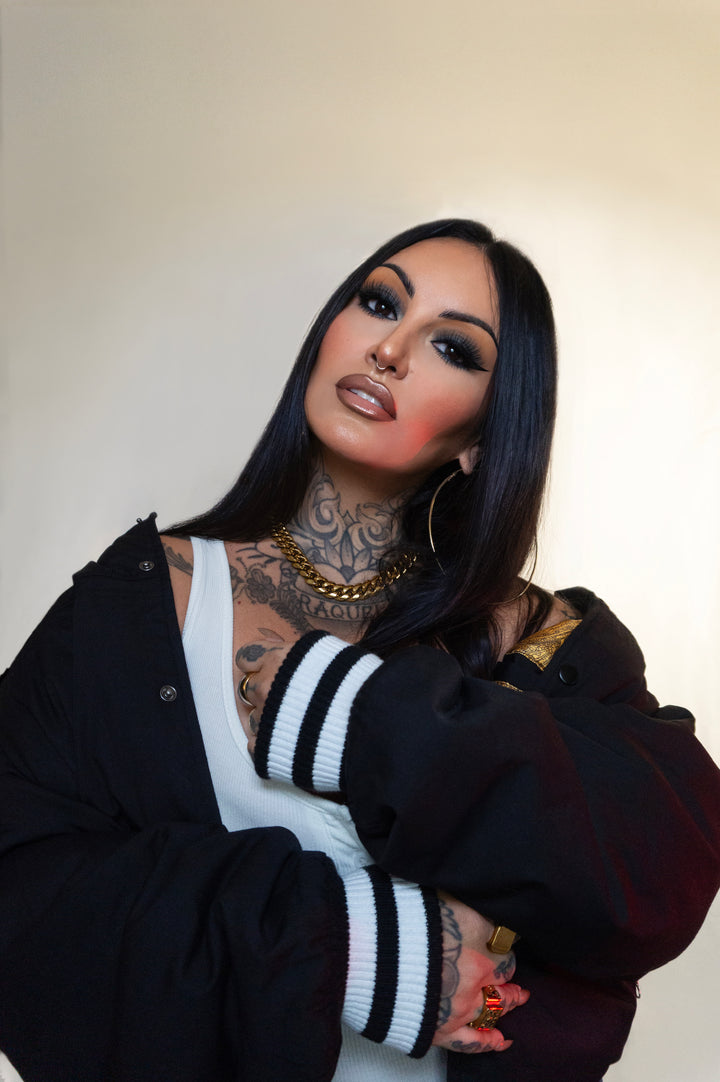 Person wearing a black jacket with white stripes, tattoos, and jewelry against a plain background