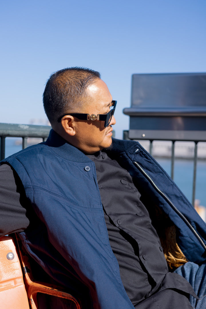 A man in profile wearing black sunglasses with gold-toned frames, a navy quilted vest over a black shirt, seated outdoors under a clear blue sky.