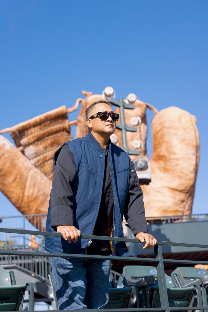 A man wearing a navy blue zip-up vest over a black shirt and jeans stands on an outdoor deck in front of a large bronze sculpture under a clear blue sky.