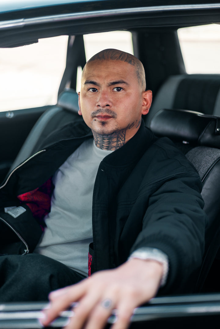 A man with a shaved head and neck tattoos sits in a black car interior, wearing a black jacket over a light gray t-shirt.
