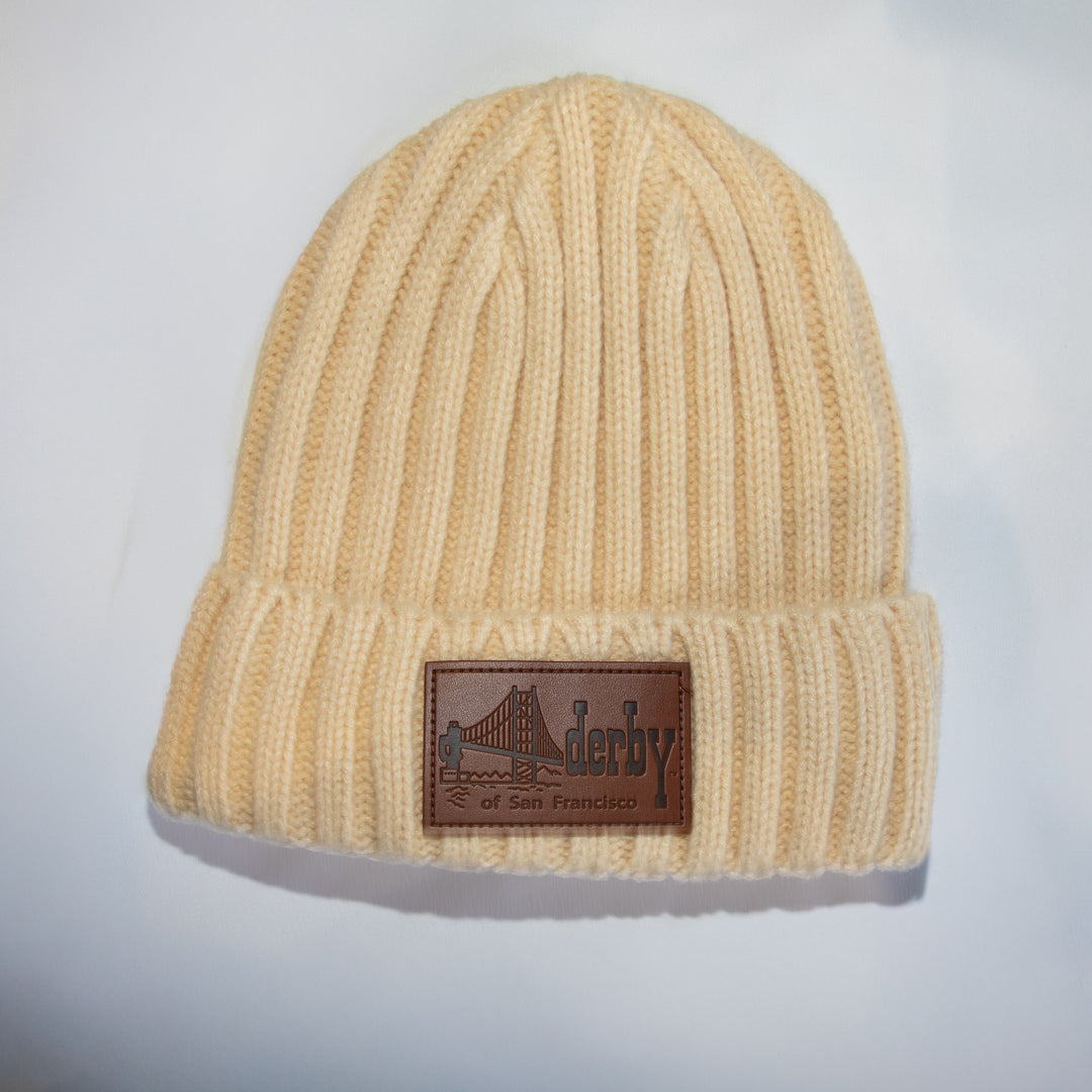 A cream-colored ribbed knit beanie hat featuring a brown rectangular leather patch on the front cuff with a bridge graphic and the text &