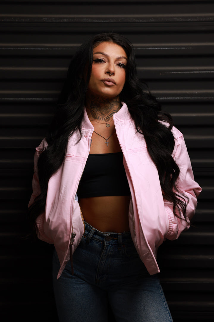 A woman with long black hair stands against a dark corrugated metal background, wearing a light pink bomber jacket over a black crop top and blue jeans.