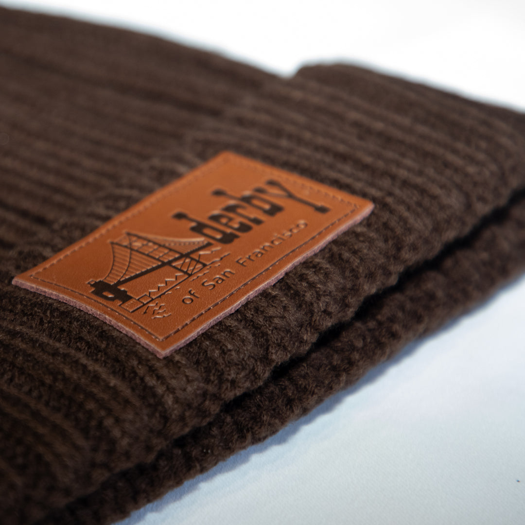 A close-up of a dark brown ribbed knit beanie with a tan leather patch on the side featuring the embroidered brand name &