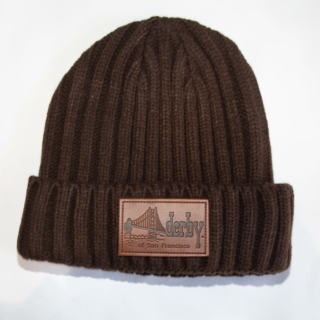 A dark brown ribbed knit beanie hat with a folded cuff, featuring a rectangular brown leather patch on the front with an embossed Golden Gate Bridge design and the text &