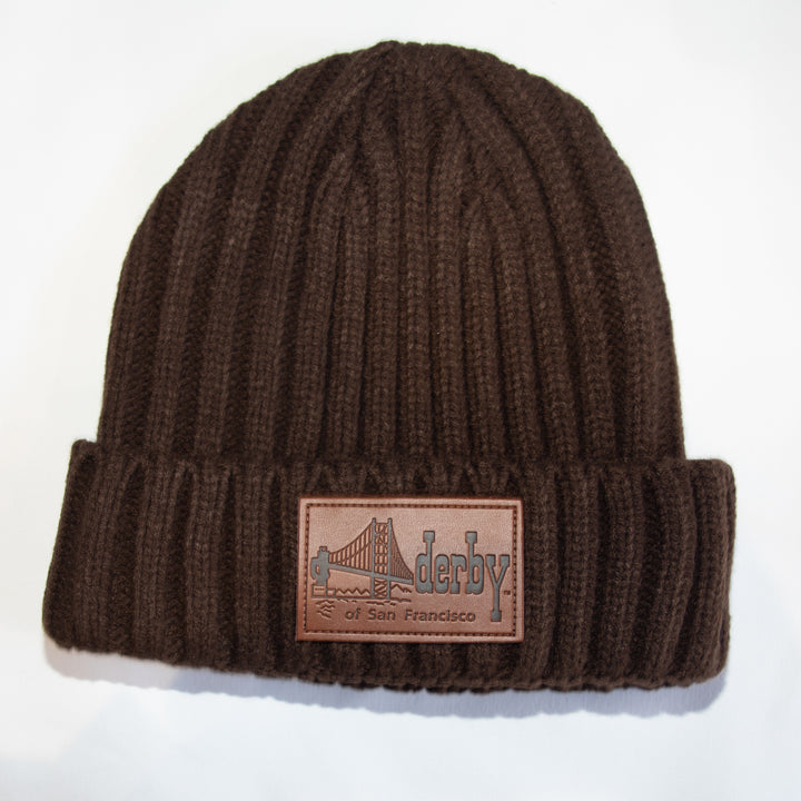 A dark brown ribbed knit beanie hat with a folded cuff, featuring a rectangular brown leather patch on the front with an embossed Golden Gate Bridge design and the text &