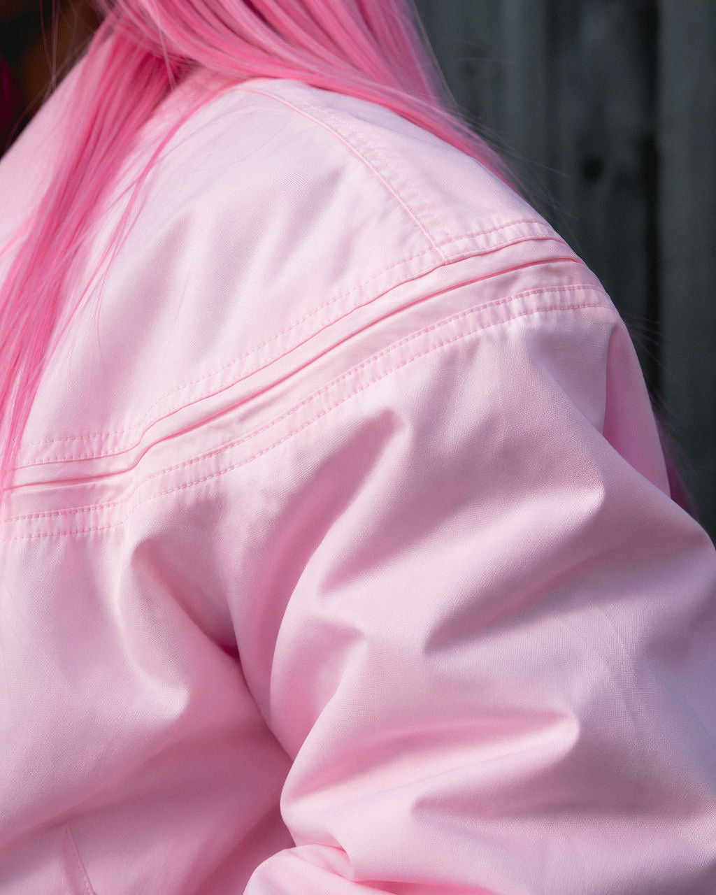 Close-up of a person wearing a light pink jacket, showing the shoulder and upper back with bright pink hair against a dark gray background.