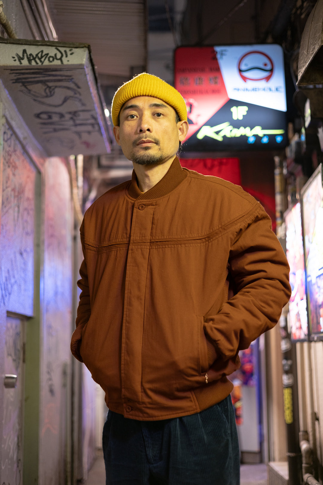 Man wearing a brown jacket and yellow beanie standing in an urban alleyway with neon signs.
