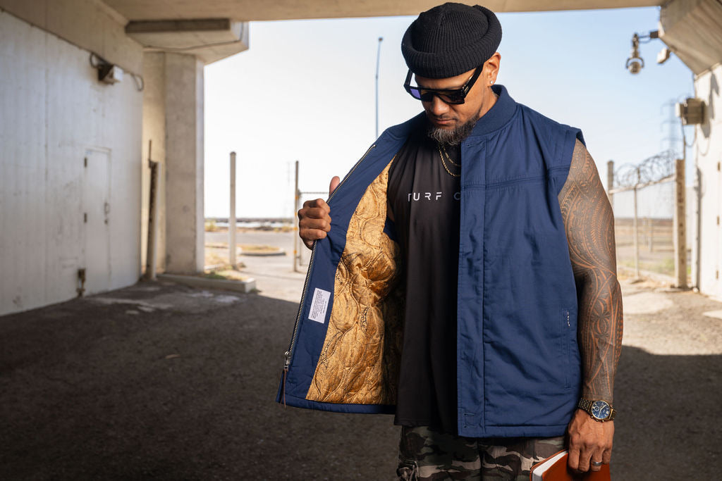 A man wearing a navy blue sleeveless vest over a black t-shirt stands in an outdoor concrete structure, holding the vest open to show a gold patterned lining.